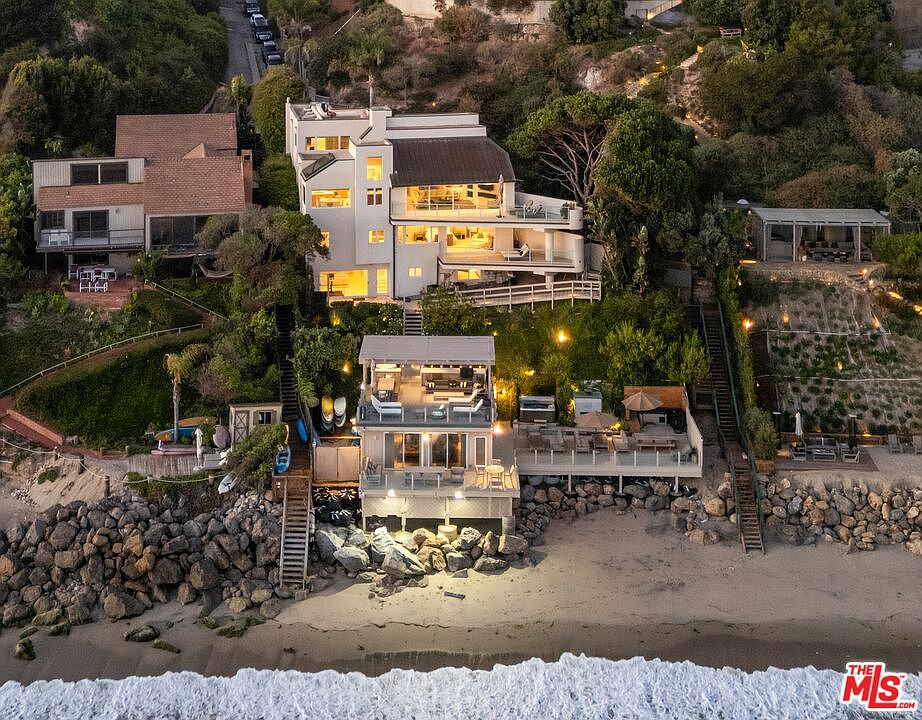 This aerial shot showcases a multi-level beachfront property with multiple decks and outdoor living spaces. The architecture features a modern design with clean lines and large windows, offering stunning ocean views. The property includes direct beach access and is surrounded by lush landscaping, creating a private and luxurious coastal retreat.