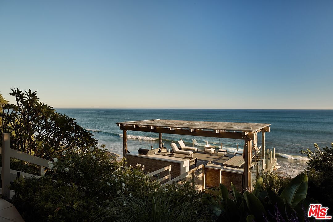 This image showcases a luxurious outdoor patio/deck area overlooking the ocean. The space features a covered structure with lounge chairs, a built-in grill, and glass railings, providing unobstructed views of the water. Lush greenery surrounds the deck, creating a private and serene atmosphere, perfect for relaxation and entertaining.