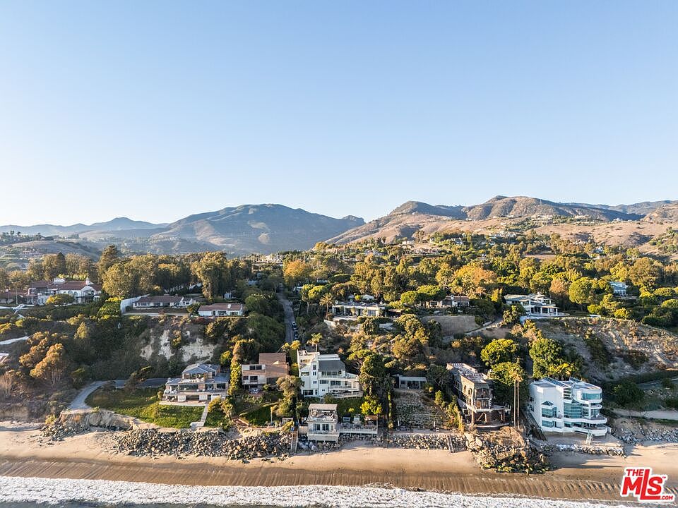 This aerial view showcases a collection of luxury homes nestled along a picturesque coastline, with sandy beaches meeting the ocean. The properties are surrounded by lush greenery and rolling hills, creating a serene and exclusive atmosphere. The clear blue sky enhances the overall appeal, highlighting the desirable location and upscale living.