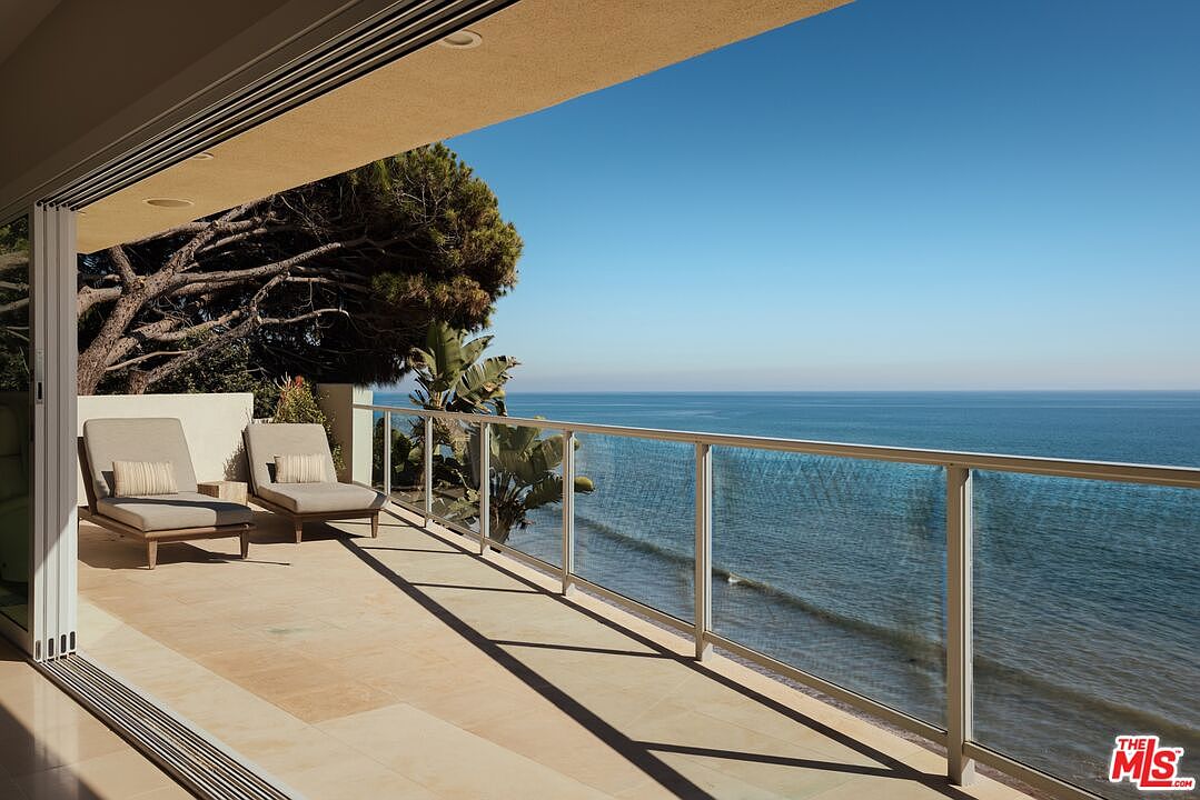 This image showcases a luxurious balcony with stunning ocean views. The balcony features a modern glass railing, comfortable lounge chairs, and a tiled floor. The open doors create a seamless transition between indoor and outdoor living, emphasizing the property's desirable location and upscale design.