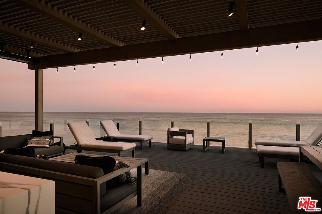This image showcases a luxurious outdoor deck or patio area with stunning ocean views at sunset. The space is furnished with modern lounge chairs, sofas, and tables, creating a relaxing and inviting atmosphere. A wooden pergola with string lights adds a touch of elegance, while the glass railing ensures unobstructed views of the ocean.