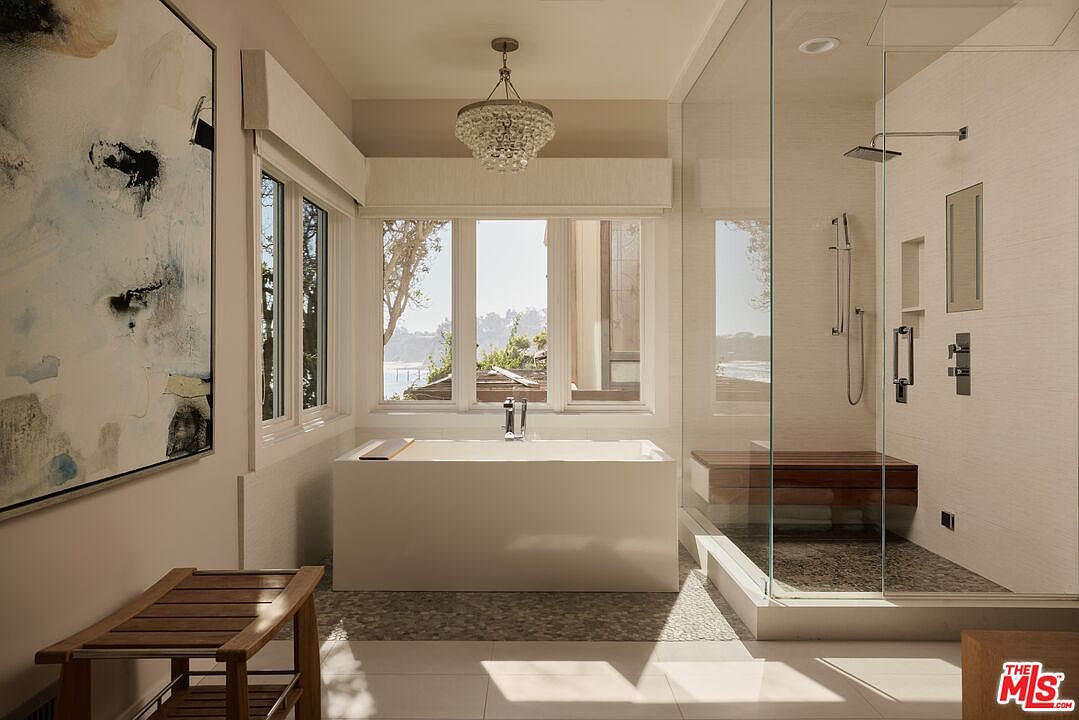 This is a bright and luxurious primary bathroom featuring a modern freestanding bathtub positioned in front of a large window with a view. A glass-enclosed shower with a built-in bench adds to the spa-like atmosphere, while a decorative chandelier provides elegant lighting. The color palette is neutral, creating a serene and sophisticated space.