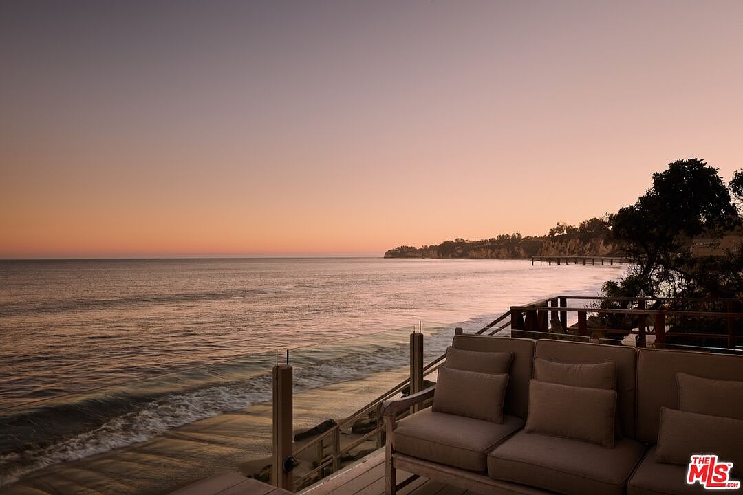 This image showcases a serene patio or deck overlooking the ocean at sunset. Comfortable outdoor seating is arranged on the wooden deck, providing an inviting space to relax and enjoy the coastal view. The scene evokes a sense of tranquility and luxury, highlighting the property's prime waterfront location.