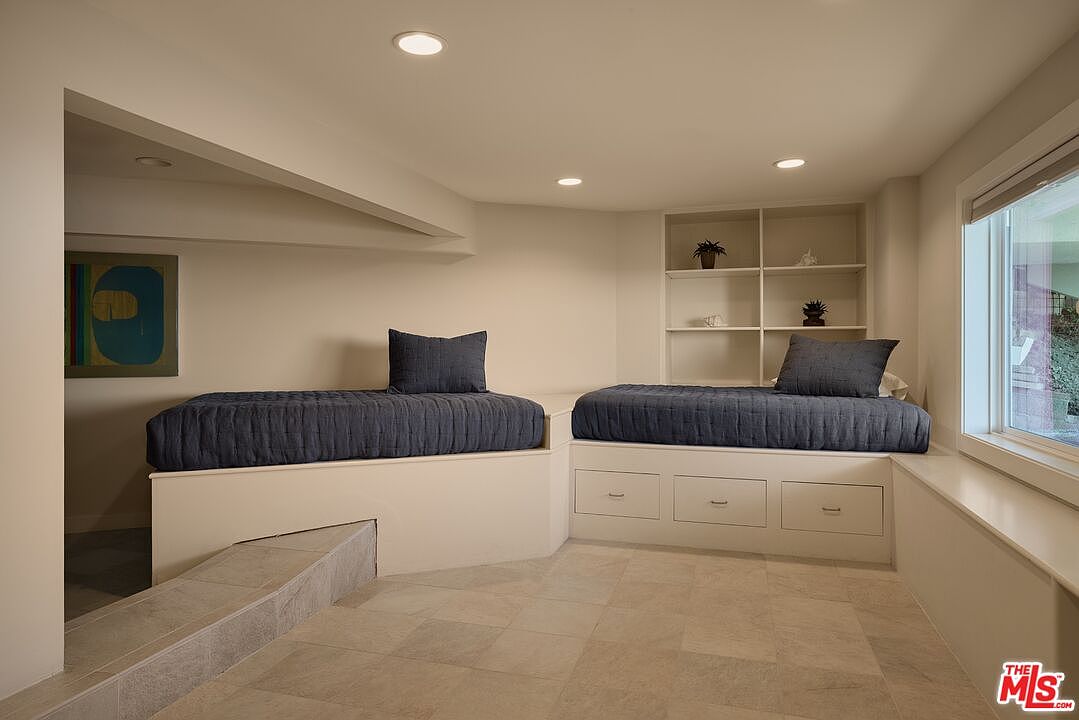 This interior shot showcases a cozy guest bedroom featuring two built-in daybeds with navy blue quilted covers and matching pillows. The room is well-lit with recessed lighting and natural light from a window, complemented by built-in shelving. The neutral color palette and tile flooring create a clean and inviting space.
