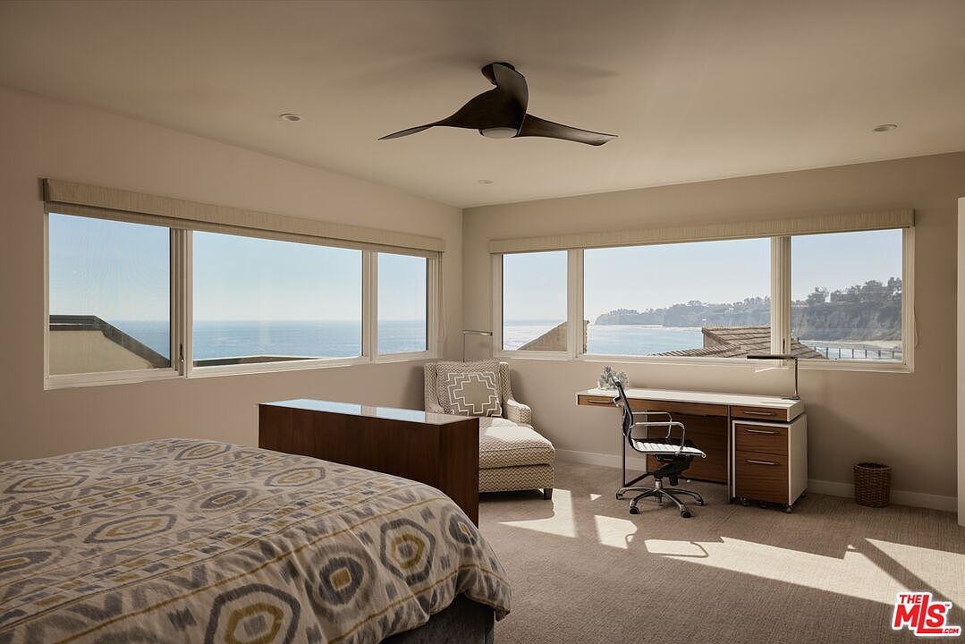 This primary bedroom features a serene ocean view through large windows, complemented by a modern ceiling fan and neutral color palette. The room includes a desk area with a rolling chair, a chaise lounge, and a bed with a patterned duvet, creating a comfortable and functional space. The overall impression is bright, airy, and relaxing.