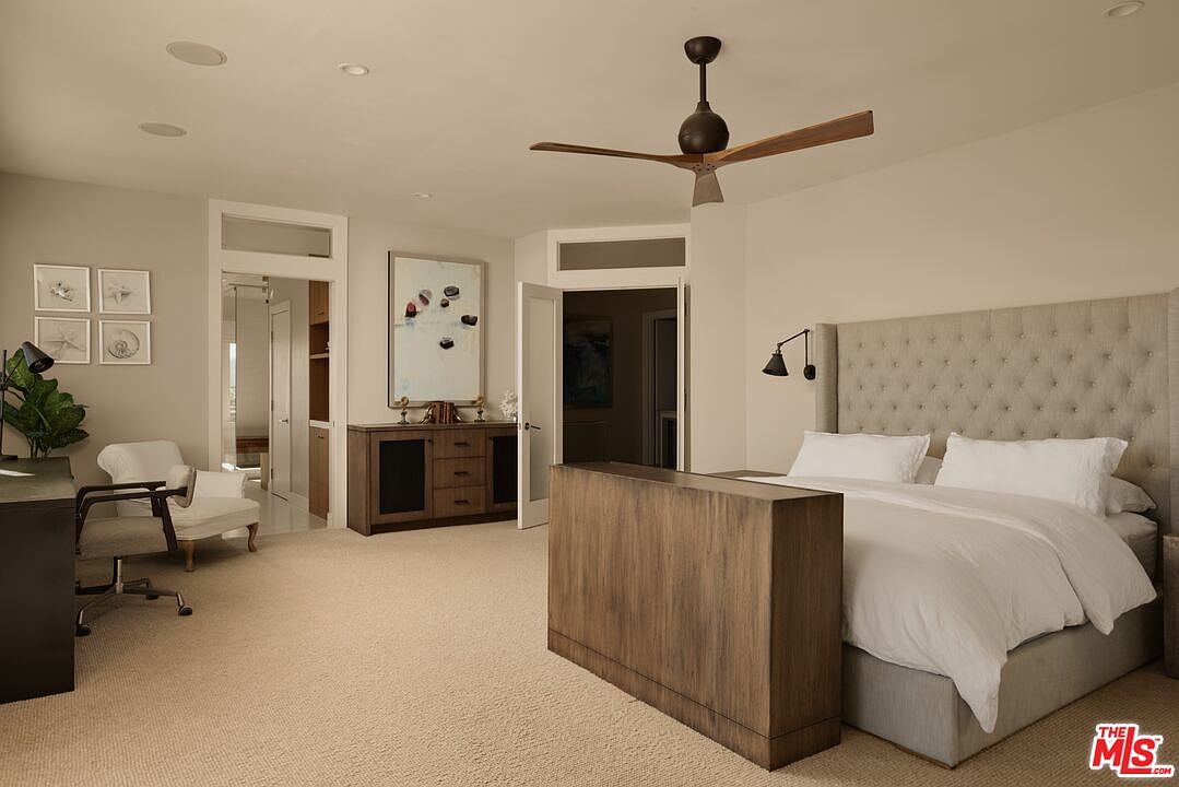 This is a primary bedroom featuring a large upholstered headboard, a bed with white linens, and a wooden console at the foot of the bed. The room includes a ceiling fan, recessed lighting, and a desk area with a chair. There is also a doorway leading to another room or closet. The perspective is from the doorway, looking into the bedroom.