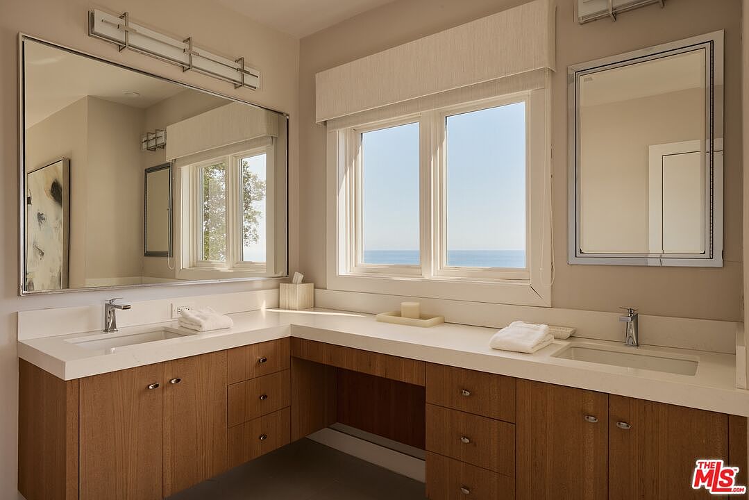 This is a well-lit primary bathroom featuring a double vanity with light wood cabinetry and white countertops. Two mirrors hang above the sinks, and a large window provides natural light and a view of the ocean. The overall design is clean and modern, creating a serene and luxurious atmosphere.