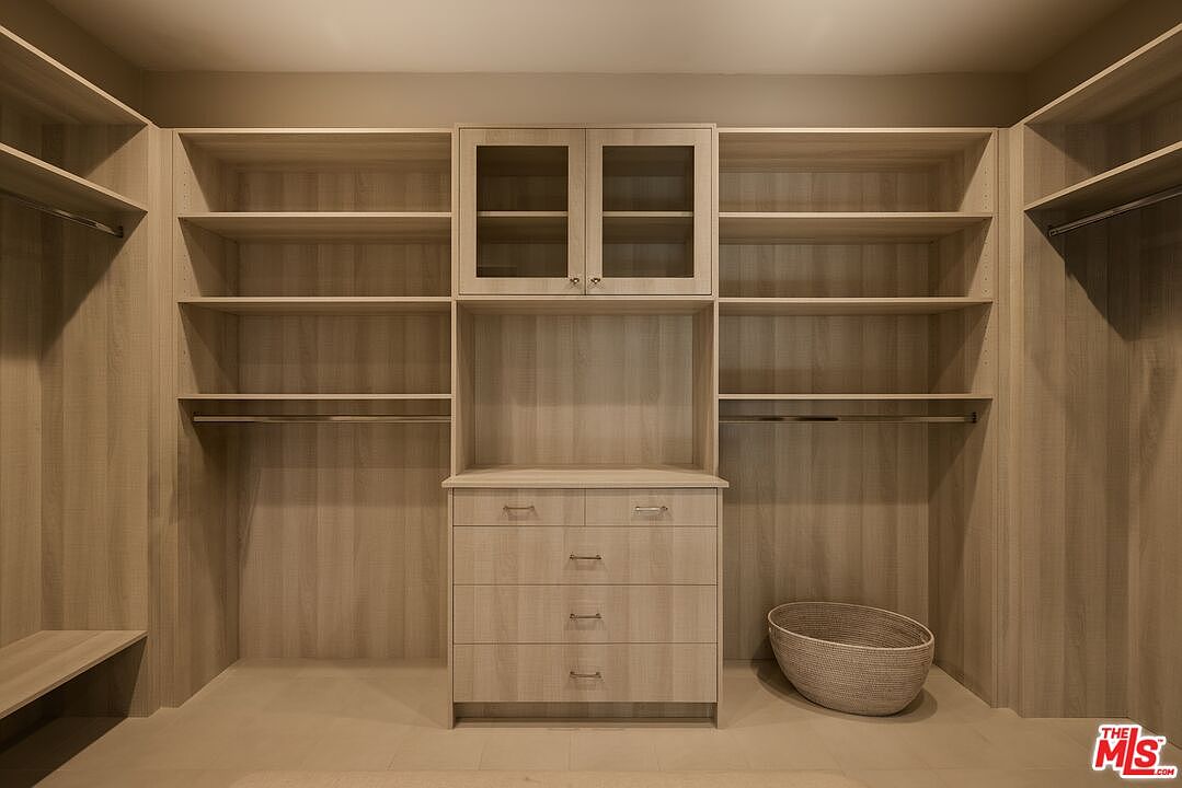 This is a well-organized walk-in closet featuring custom shelving and drawers. The closet is designed with light wood finishes and includes both open shelving and closed storage with glass-front cabinets. A woven basket sits on the floor, adding a touch of texture to the space.