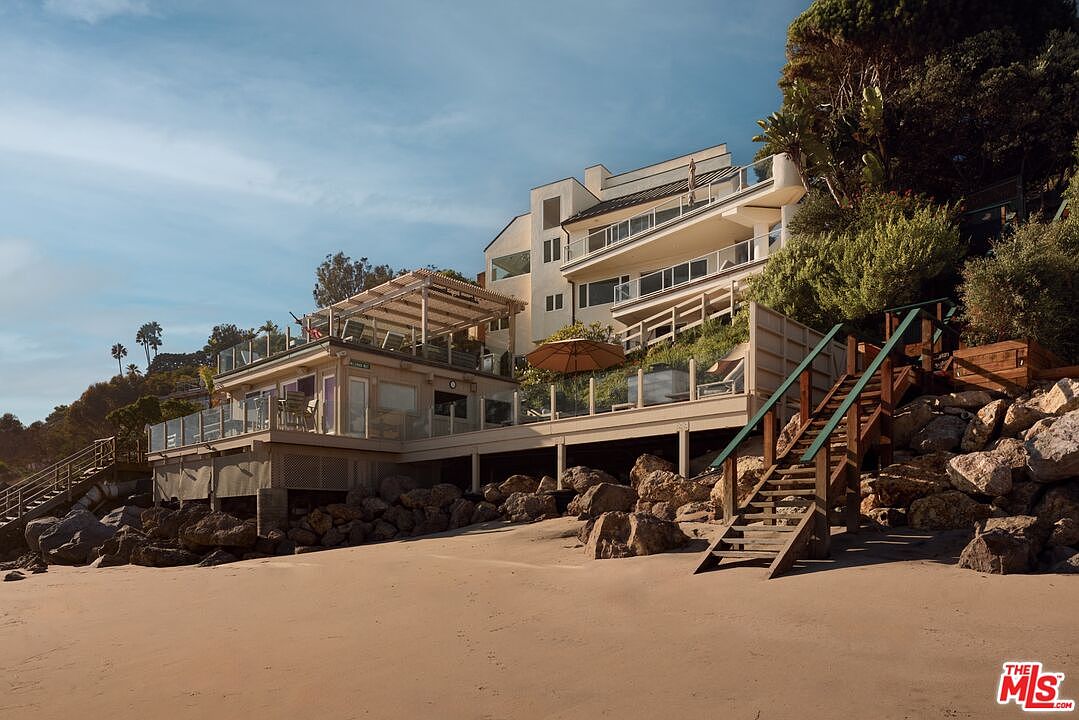 This exterior shot showcases the rear of a multi-level beachfront property. The architecture features a blend of modern and coastal styles, with multiple decks and balconies offering ocean views. Wooden staircases provide access to the beach, and the property is nestled among rocks and vegetation, creating a private and scenic setting.