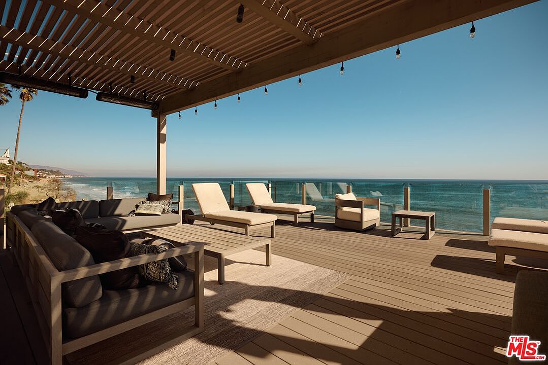 This image showcases a luxurious outdoor patio or deck area with stunning ocean views. The space is furnished with comfortable seating, including a large sofa and lounge chairs, all under a stylish pergola with hanging lights. The deck features a glass railing to maximize the view, creating an inviting and relaxing atmosphere perfect for enjoying the coastal scenery.