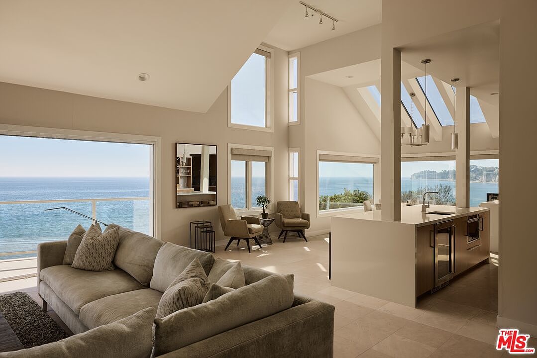 This interior shot showcases a bright and airy living space with an open floor plan that seamlessly integrates with the kitchen area. The living room features a comfortable-looking sofa with decorative pillows, while large windows offer stunning ocean views. The kitchen island is visible in the background, contributing to the modern and luxurious feel of the home.