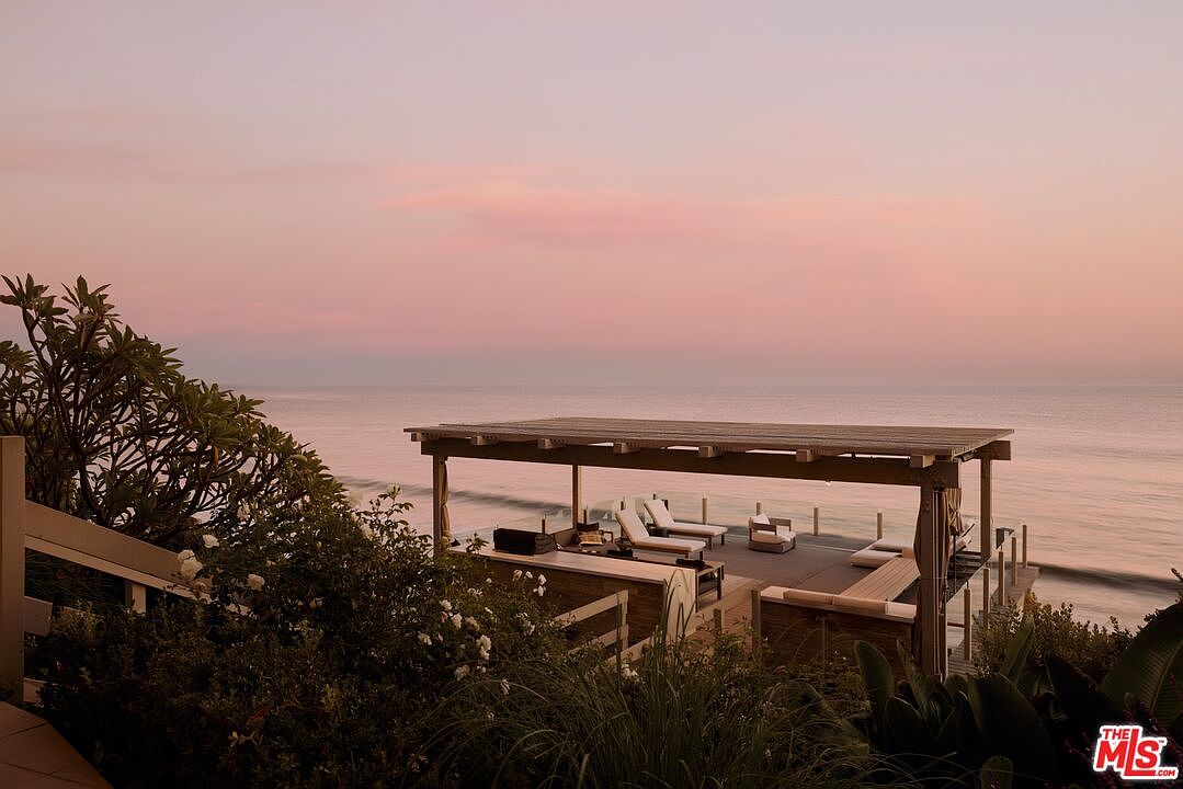 This image showcases a luxurious outdoor patio/deck/balcony overlooking the ocean at sunset. The space features a covered pergola, lounge chairs, a built-in bar area, and comfortable seating, creating an inviting atmosphere for relaxation and entertainment. Lush greenery surrounds the deck, enhancing the sense of privacy and tranquility.