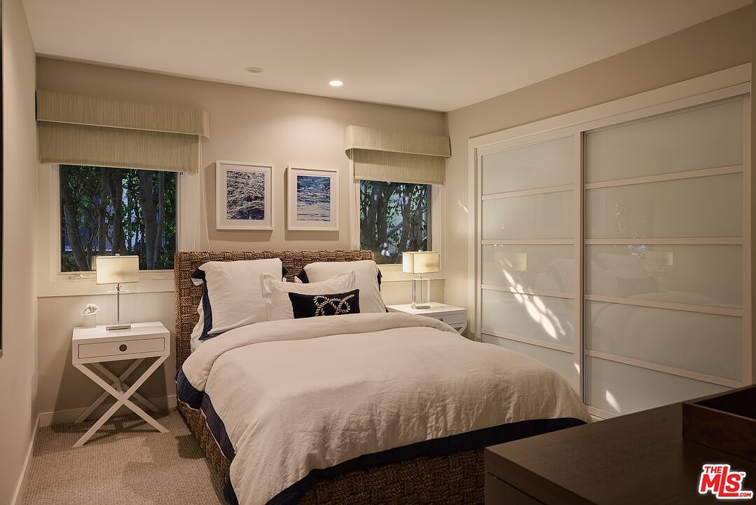 This is a cozy bedroom featuring a bed with white linens and a navy blue accent, flanked by matching nightstands and lamps. Two framed art pieces hang above the bed, and the room is illuminated by natural light filtering through the windows. A sliding door closet is on the right side of the room.