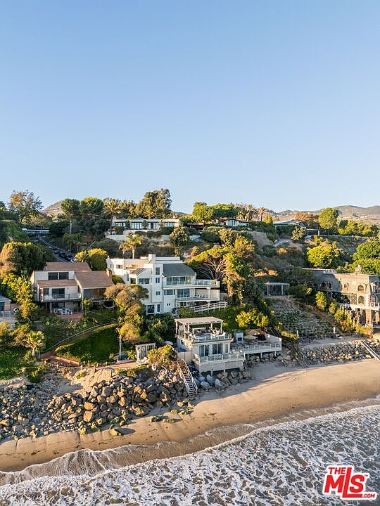 This aerial view showcases a collection of luxurious beachfront homes nestled along a picturesque coastline. The properties feature multi-level decks, modern architectural designs, and direct access to a sandy beach. The scene is bathed in warm sunlight, highlighting the natural beauty of the ocean and surrounding landscape.