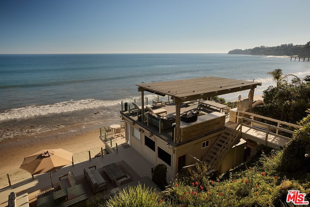 This aerial view showcases a stunning beachfront property with a multi-level deck and patio area. The house features a covered rooftop deck with lounge seating, offering panoramic ocean views. A private beach access and lush landscaping enhance the property's appeal, creating a luxurious coastal retreat.