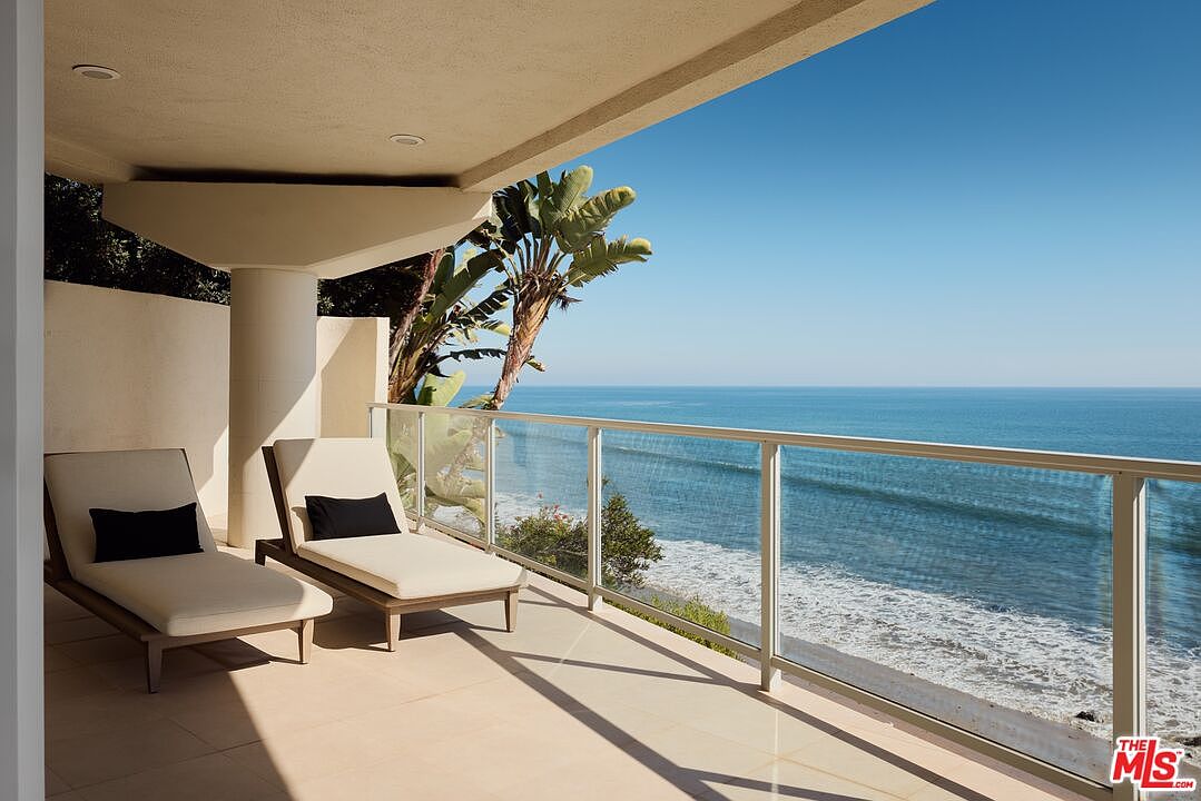 This image showcases a luxurious balcony with stunning ocean views. Two chaise lounges with black pillows invite relaxation, while a modern glass railing ensures unobstructed scenery. The neutral-toned tile flooring and architectural details create an elegant and serene outdoor living space.