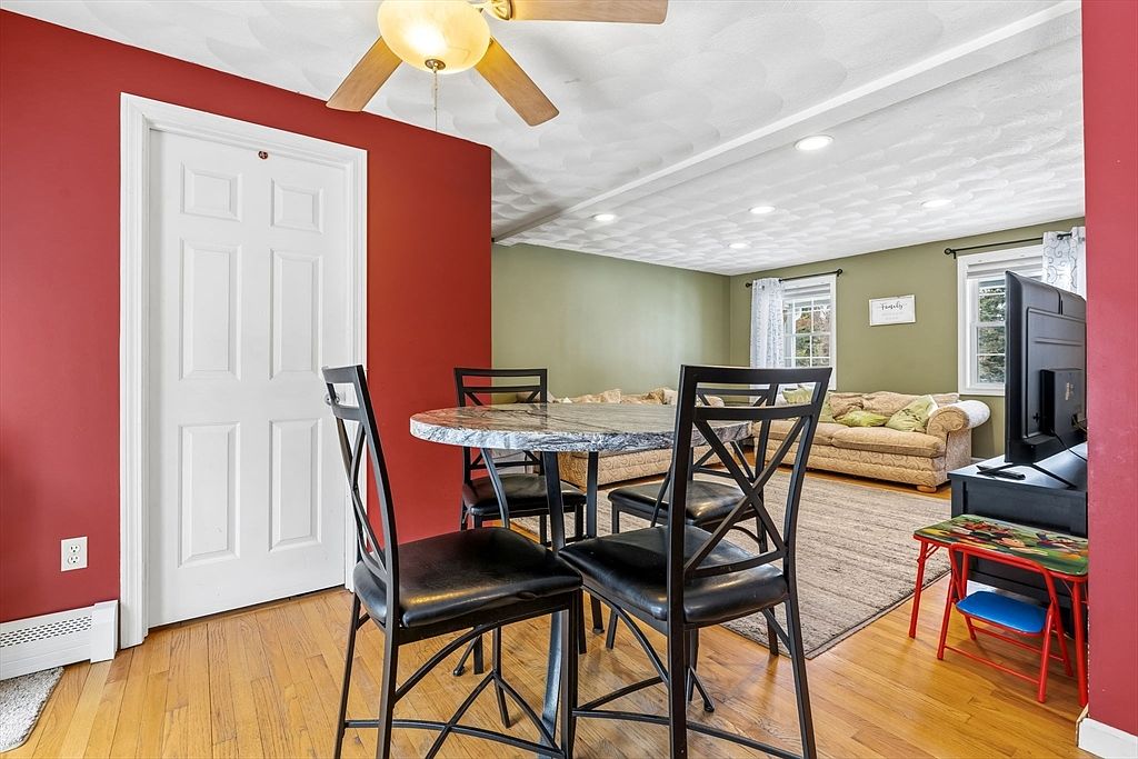This interior shot showcases a dining area with a round marble-top table surrounded by four black chairs. The room features a combination of red and green walls, hardwood flooring, and a view into a living room area with a beige sofa and a television. A white door is visible on the left, adding to the room's character.