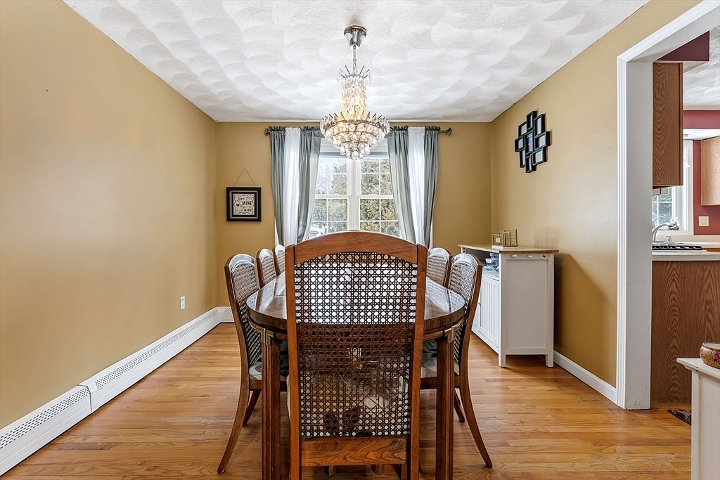 This is a dining room featuring a round wooden table with cane-backed chairs. A crystal chandelier hangs above the table, providing elegant lighting. The walls are painted a warm yellow, and a window with curtains allows natural light to filter into the room. A white sideboard sits against one wall, adding storage and display space.