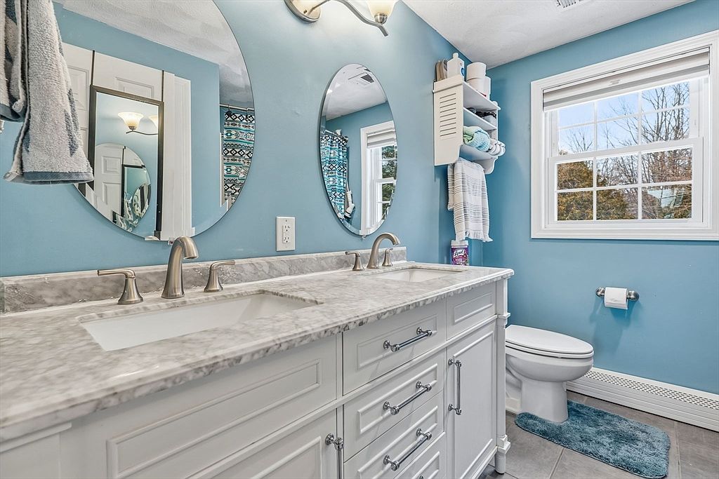 This is a well-lit bathroom featuring a double vanity with a marble countertop and white cabinetry. Two oval mirrors hang above the sinks, reflecting the blue walls and a window that provides natural light. A toilet is visible to the right, and the overall impression is clean and inviting.