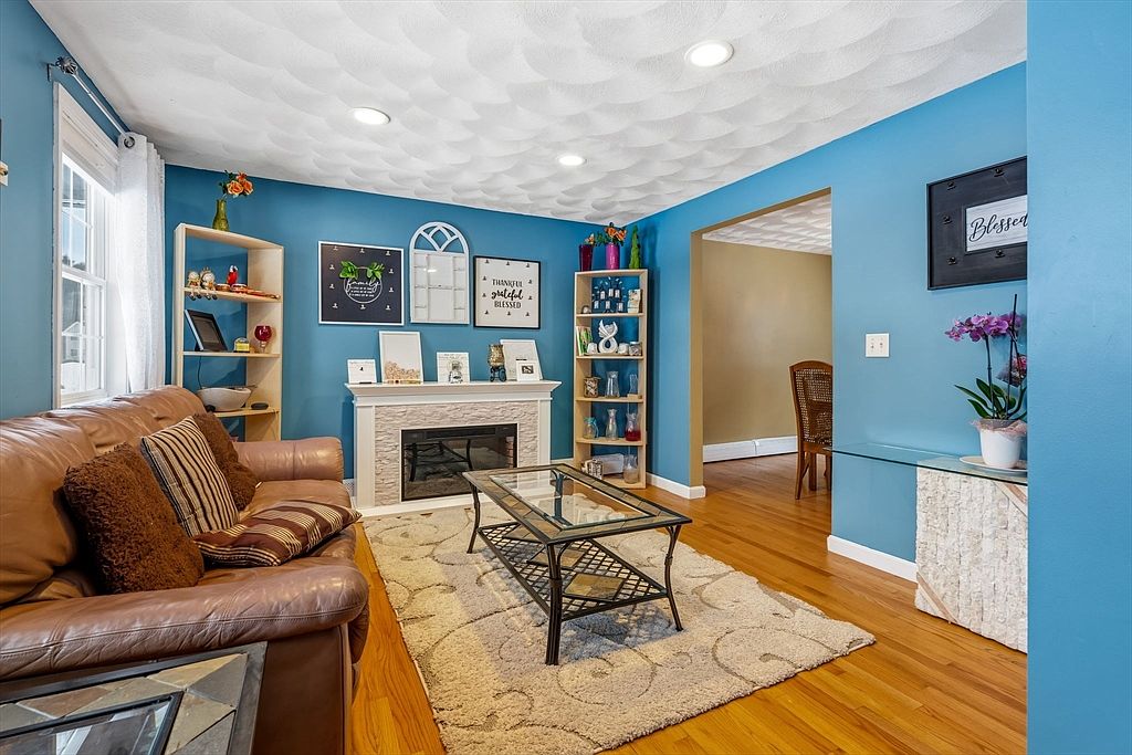 This is a cozy living room featuring blue walls, hardwood floors, and a textured ceiling. A brown leather sofa is adorned with decorative pillows, and a glass-topped coffee table sits on a patterned rug. A fireplace with decorative items and shelving units on either side add to the room's charm, creating a warm and inviting atmosphere.