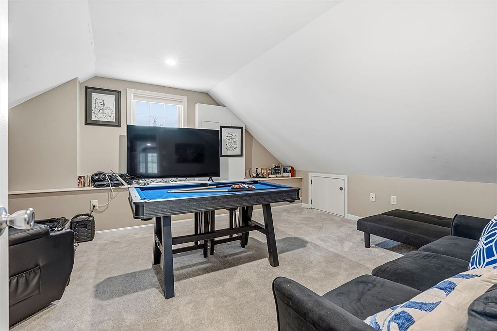This is an interior shot of an attic space, likely converted into a recreational room. The room features a pool table, a large television, and a comfortable-looking sofa. The walls are painted in neutral tones, and the carpet is a light gray, creating a cozy and inviting atmosphere.