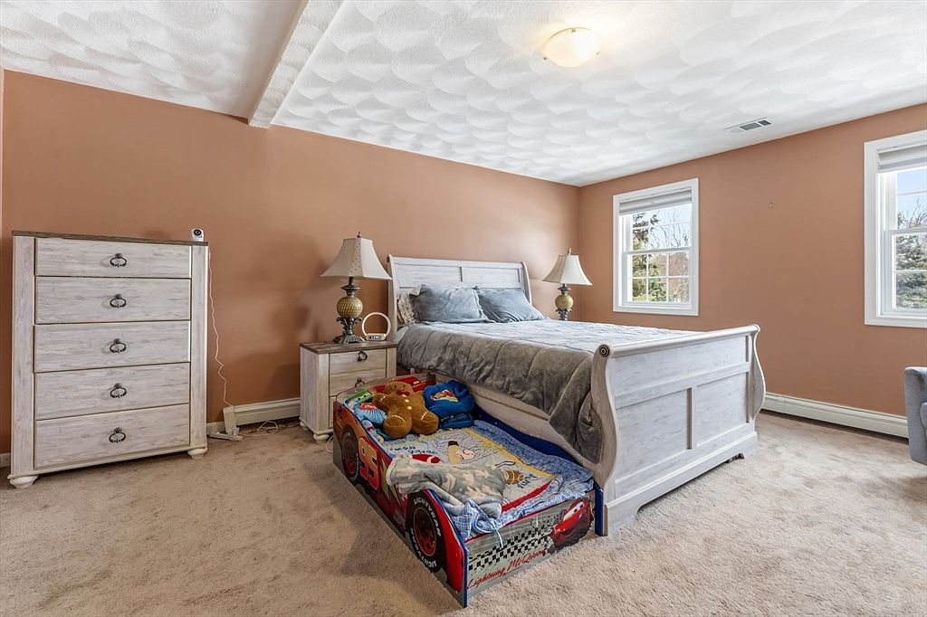 This is a cozy bedroom featuring a white wooden bed with a trundle bed underneath, a matching dresser, and a nightstand with a lamp. The walls are painted in a warm terracotta color, and the room is carpeted. Natural light streams in through two windows, creating a bright and inviting space.