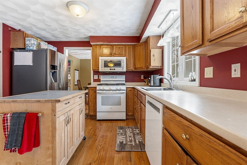 This is a warm and inviting kitchen with wood cabinetry, stainless steel appliances, and a kitchen island. The walls are painted a deep red, contrasting with the light countertops and wood flooring. Natural light streams in from a window above the sink, creating a cozy atmosphere.