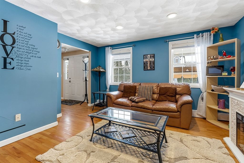 This is an interior shot of a living room featuring a brown leather sofa with throw pillows, a glass-topped coffee table on a patterned rug, and hardwood floors. The walls are painted a deep blue, and there are two windows with white curtains, along with a bookshelf and a decorative fireplace. The room has a cozy and inviting atmosphere.