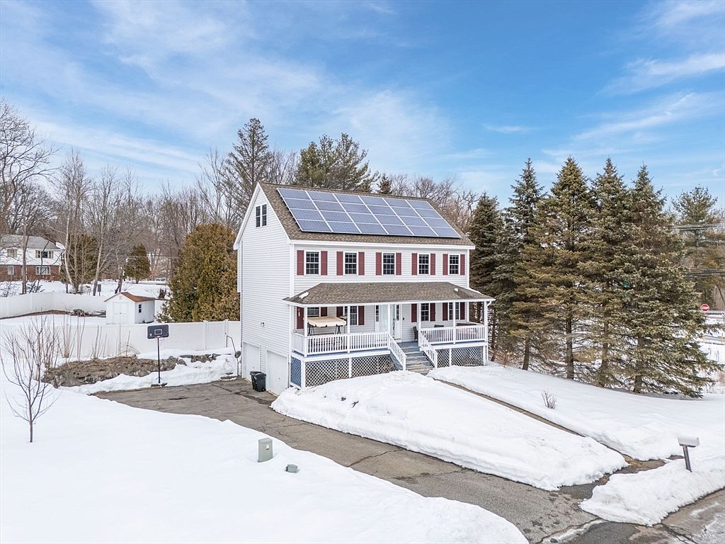 This is a front exterior view of a two-story house with a covered porch. The house has white siding, red shutters, and solar panels on the roof. The front yard is partially covered in snow, and there is a driveway leading up to the house.