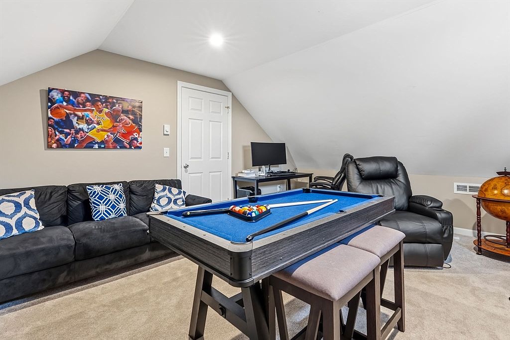 This is an interior shot of a finished attic space, featuring a pool table as a central element. The room includes a dark-colored sofa with decorative pillows, a black leather recliner, and a desk with a computer. A basketball-themed artwork hangs on the wall, and the space is illuminated by a ceiling light, creating a cozy and recreational atmosphere.
