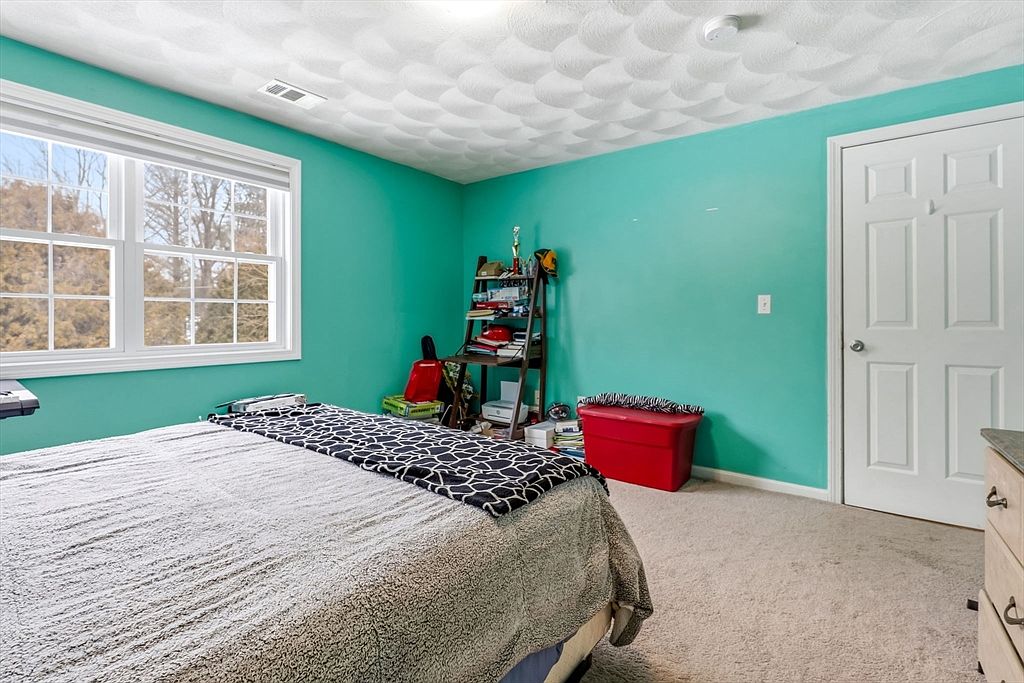 This is a bedroom with teal walls and a textured white ceiling. A bed with a gray and black patterned blanket is the focal point, positioned near a window that provides natural light. A ladder shelf with various items and a red storage bin are against the wall, and a white door is visible on the right side of the frame.