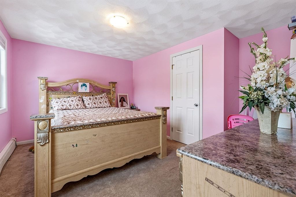 This is a bedroom with pink walls and carpeted floors. The room features a wooden bed with decorative accents, a white door, and a table with a granite top. The overall impression is a cozy, if somewhat unusually colored, space.