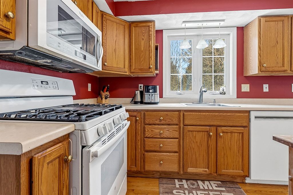 This is a kitchen with red walls and wooden cabinets. A white stove and microwave are visible on the left, and a window above the sink provides natural light. The kitchen has a warm, traditional feel.