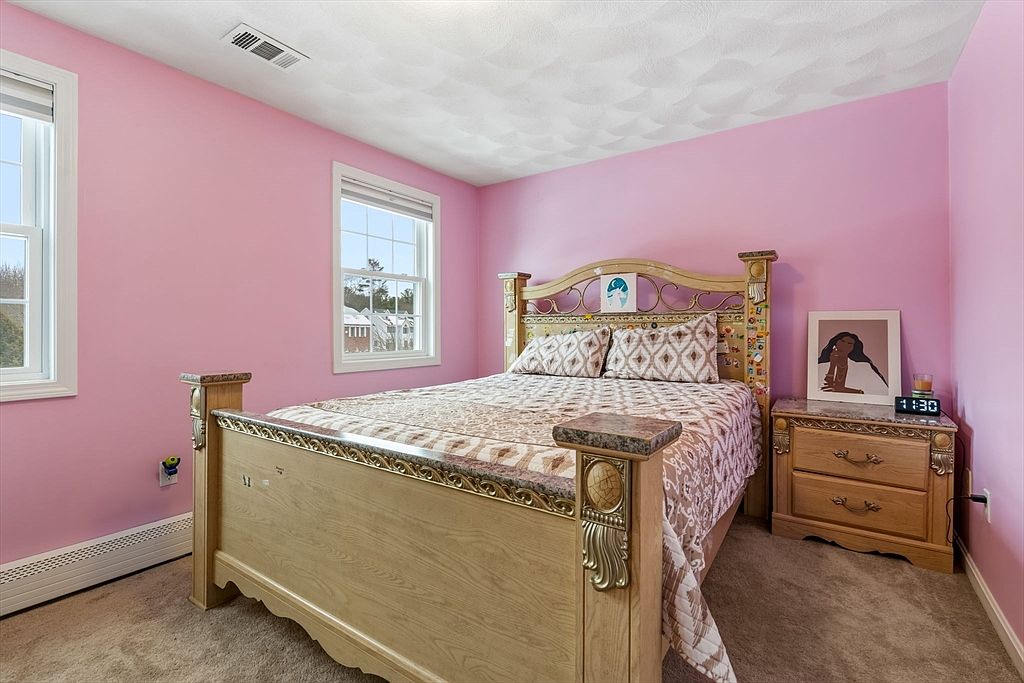 This is an interior shot of a bedroom featuring pink walls and a light wood bed with ornate detailing. The bed is neatly made with patterned bedding and pillows. A matching nightstand with a digital clock and framed artwork sits beside the bed, and natural light streams in through two windows, creating a bright and inviting atmosphere.