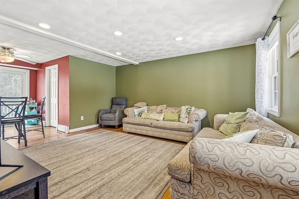 This is an interior shot of a living room featuring two patterned sofas, a gray armchair, and a neutral-toned rug. The walls are painted in a combination of olive green and red, and the ceiling has a textured pattern with recessed lighting. The room appears cozy and inviting, with a mix of traditional and contemporary elements.