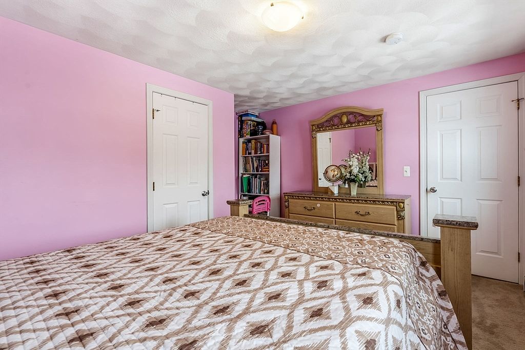 This is a cozy bedroom with pink walls and a patterned bedspread. The room features a wooden dresser with a large mirror, a built-in bookshelf, and two white doors, one possibly leading to a closet or bathroom. The overall impression is a comfortable and inviting space.