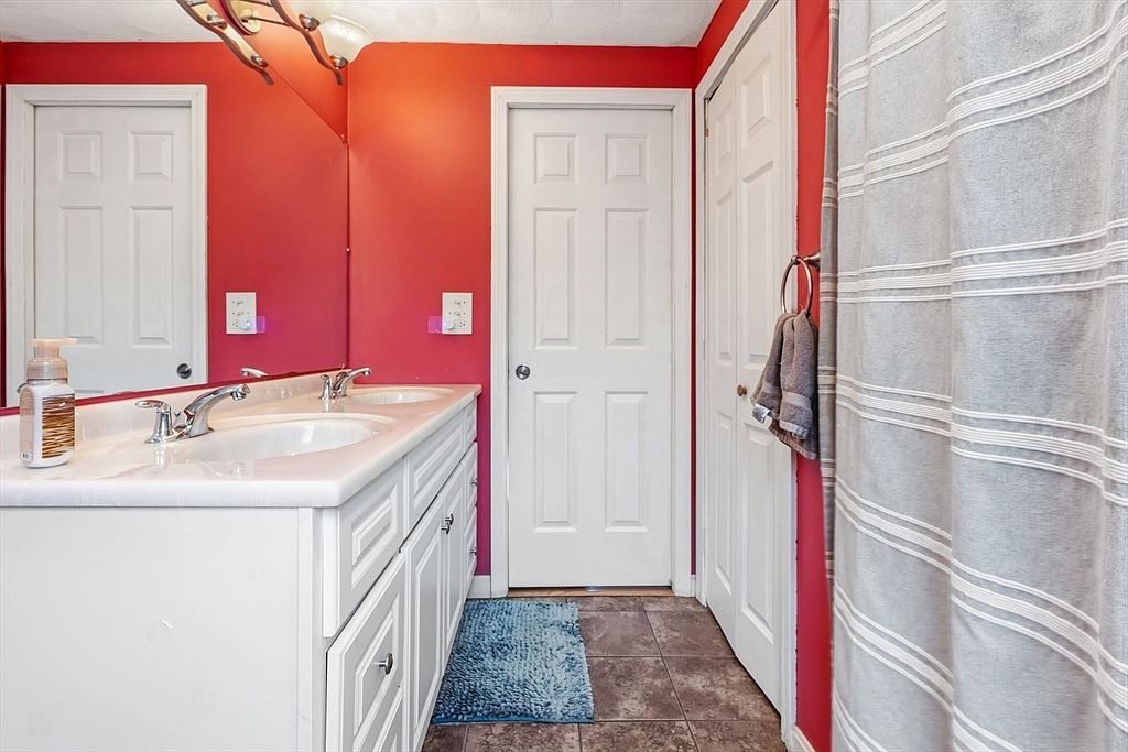 This bathroom features a double sink vanity with white cabinets and a light-colored countertop. The walls are painted a vibrant red, contrasting with the white doors and trim. A blue rug adds a pop of color to the brown tiled floor, and a striped shower curtain is partially visible on the right.