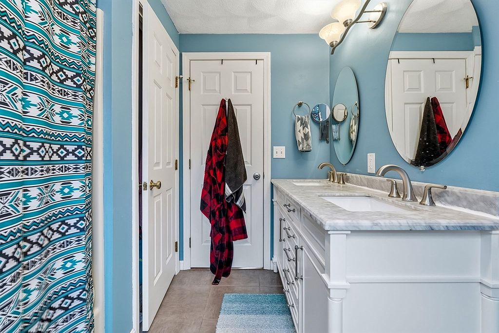 This is a well-lit bathroom featuring a double vanity with a white cabinet base and a marble countertop. Two oval mirrors hang above the sinks, complemented by modern light fixtures. A red and black plaid towel hangs on the door, adding a pop of color to the blue walls and neutral tile flooring.