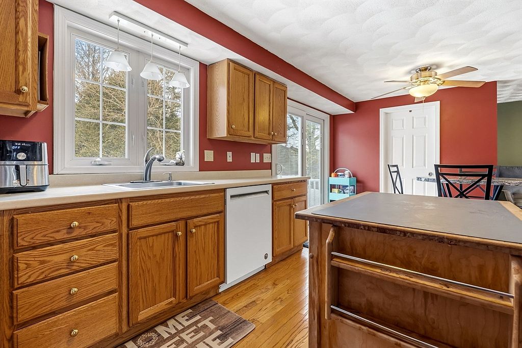 This is a warm and inviting kitchen with oak cabinetry, a light countertop, and a stainless steel sink under a window that provides natural light. The kitchen features a white dishwasher and a central island, with red walls adding a touch of color. The hardwood floors and ceiling fan complete the cozy atmosphere.