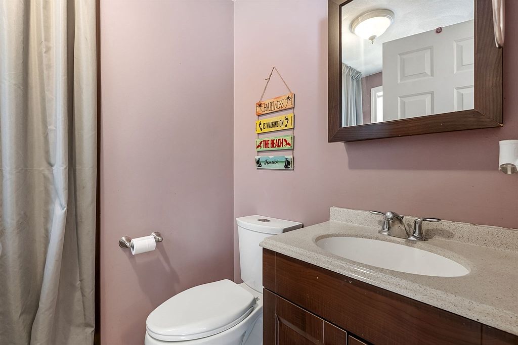 This is a cozy bathroom featuring a toilet, a vanity with a light-colored countertop and dark wood cabinetry, and a mirror. The walls are painted a soft pink, and a decorative beach-themed sign hangs on the wall. A white shower curtain is visible on the left, adding to the room's functionality.