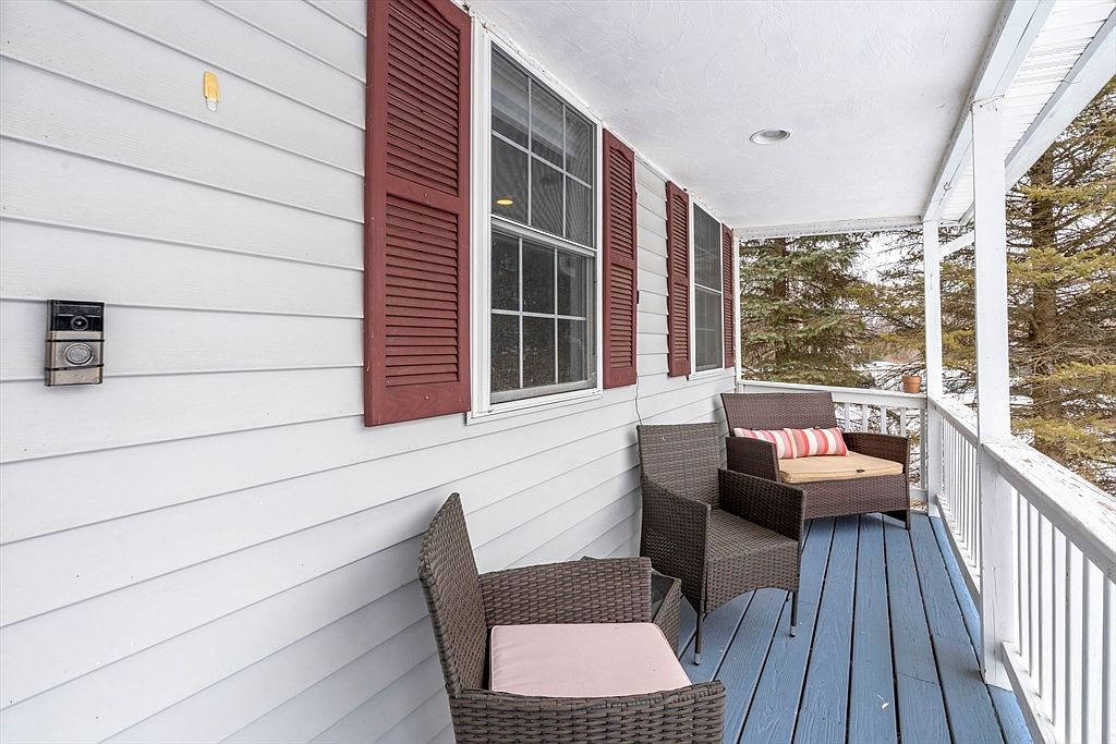This image showcases a charming porch area with two wicker chairs and a small table, creating an inviting outdoor space. The porch features a blue-painted wooden floor and white railings, complemented by the house's light gray siding and red shutters. The scene offers a cozy and relaxing atmosphere, perfect for enjoying the outdoors.