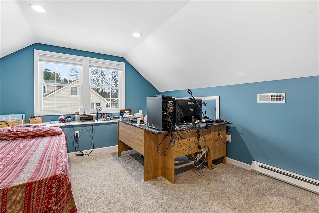 This is an interior shot of a bedroom featuring a bed with a red and white patterned comforter, a desk with a computer setup, and blue walls. The room has a sloped ceiling and a window providing natural light. The overall impression is a functional and cozy space.