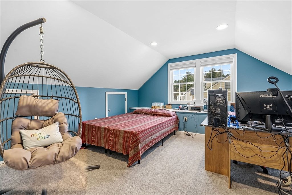 This is a cozy bedroom featuring a hanging chair, a bed with a patterned red and brown cover, and a desk with a computer setup. The walls are painted a calming blue, and the room is well-lit by natural light from the windows. The overall impression is a comfortable and functional space.