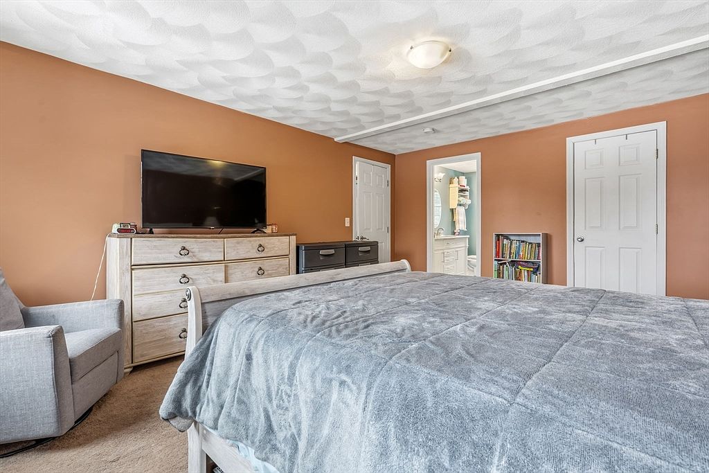 This is a primary bedroom featuring a large bed with a gray comforter, a light-colored wooden dresser with a flat-screen TV on top, and a gray armchair. The walls are painted in a warm, earthy tone, and the ceiling has a textured pattern. There are two doors visible, one leading to a bathroom and another to a closet, along with a small bookshelf.