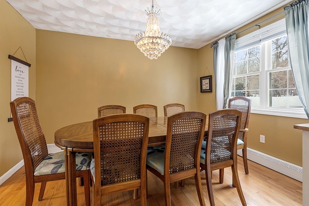 This is an interior shot of a dining room featuring a wooden oval table surrounded by six wooden chairs with woven backs and patterned cushions. A crystal chandelier hangs above the table, and natural light streams in through a window with light blue curtains. The walls are painted a warm yellow, and a decorative 'Love' sign hangs on the wall, creating a cozy and inviting atmosphere.