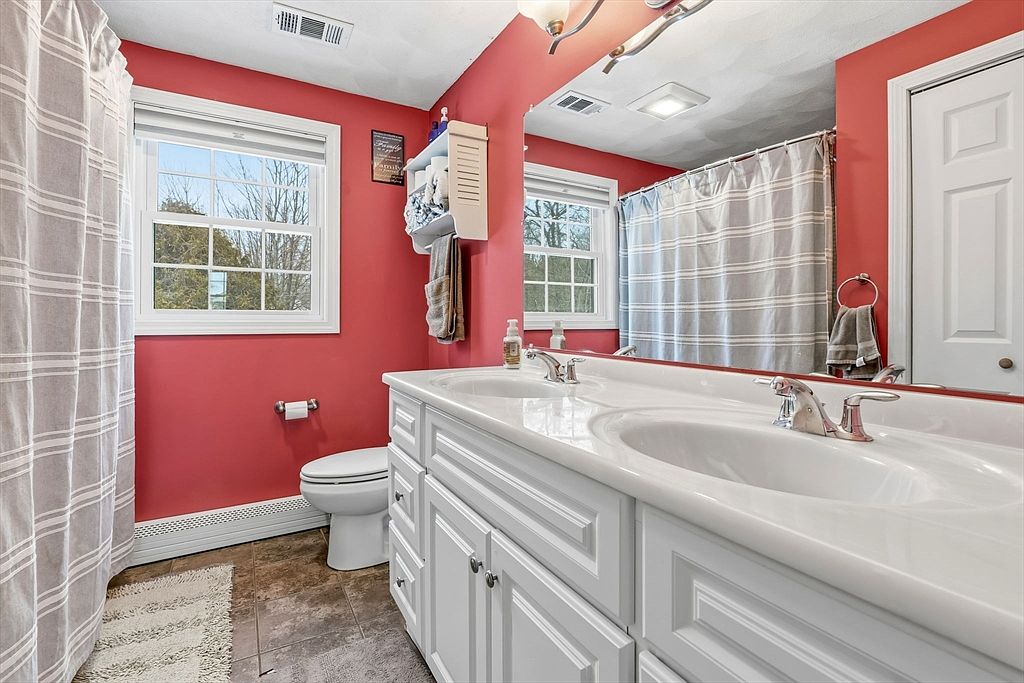 This is a bathroom featuring a double sink vanity with white cabinetry and a light countertop. The walls are painted a warm red color, and there is a window providing natural light. A shower curtain is visible, suggesting a shower/tub combo, and a toilet is positioned next to the vanity.