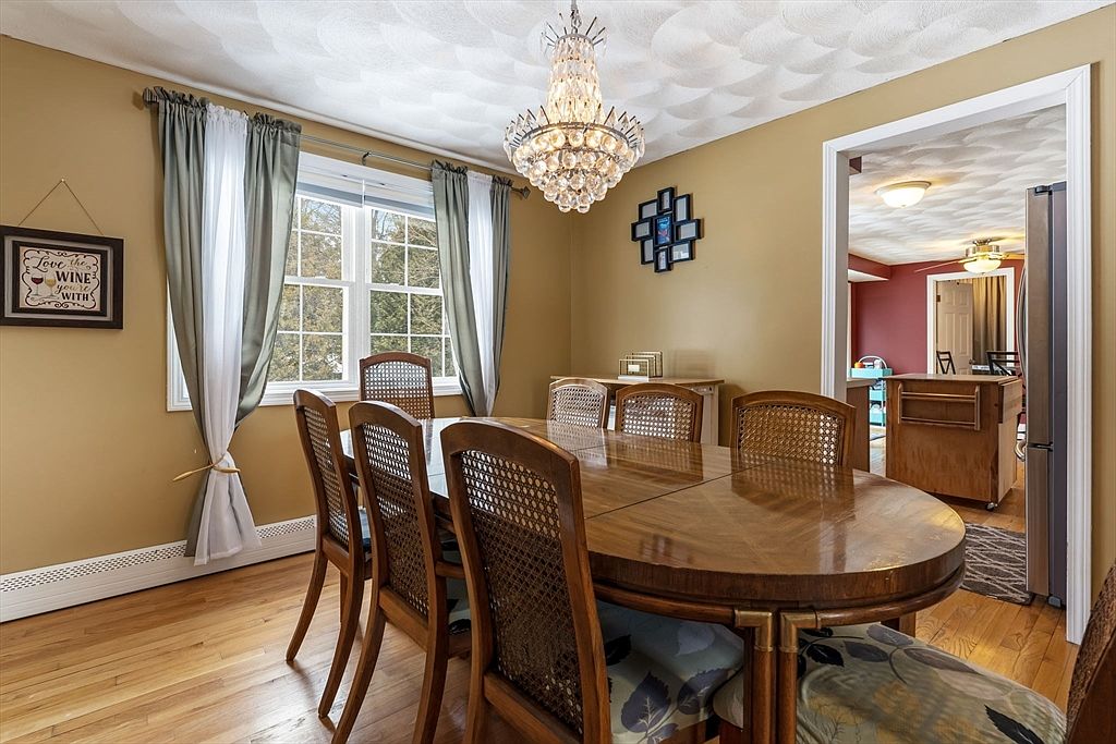 This is a dining room featuring a large wooden table with seating for eight, illuminated by a decorative chandelier. The walls are painted in a warm tone, complemented by curtains framing a window that provides natural light. The room has a traditional style with hardwood floors and a view into an adjacent room, creating an inviting atmosphere for meals and gatherings.