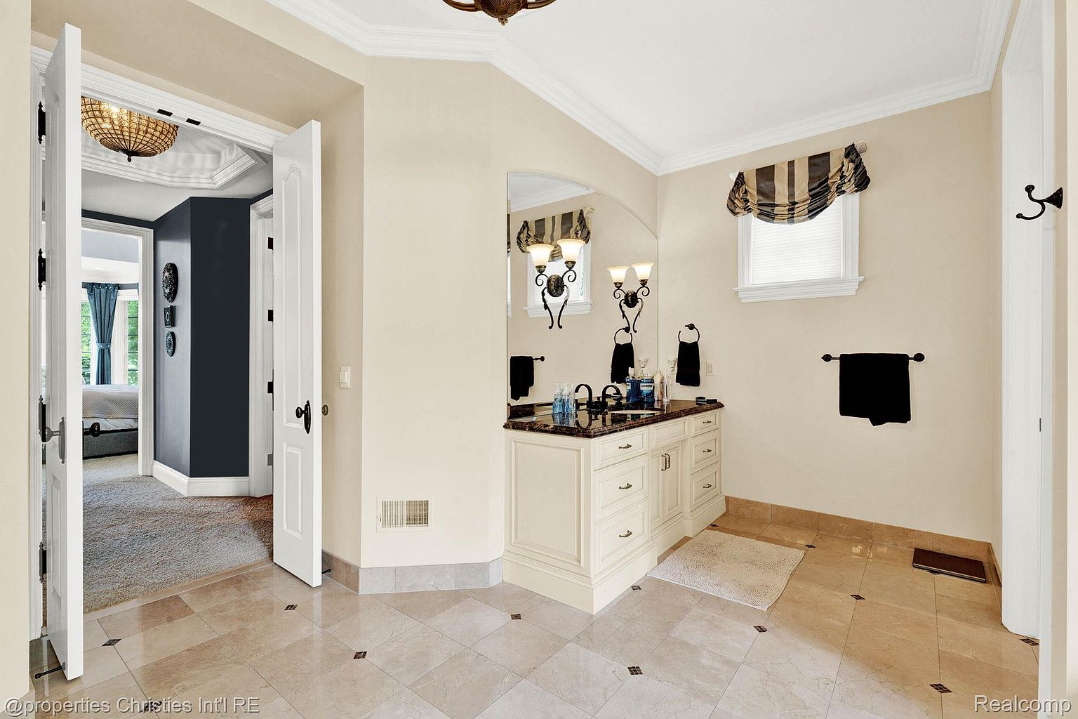 This is a well-lit primary bathroom featuring a vanity with a dark countertop and cream-colored cabinetry. A large mirror hangs above the vanity, reflecting the sconces on either side. The flooring is tiled with a diamond pattern, and a window with a decorative valance provides natural light.