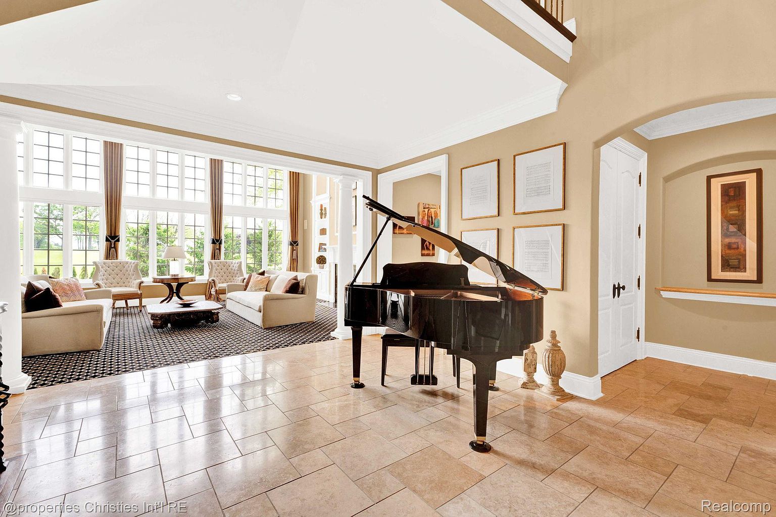 This is a spacious living room featuring a grand piano as a focal point. The room is well-lit with large windows and features elegant furniture, including sofas and armchairs, arranged around a patterned rug. The flooring is tiled, and the walls are painted in neutral tones, creating a sophisticated and inviting atmosphere.