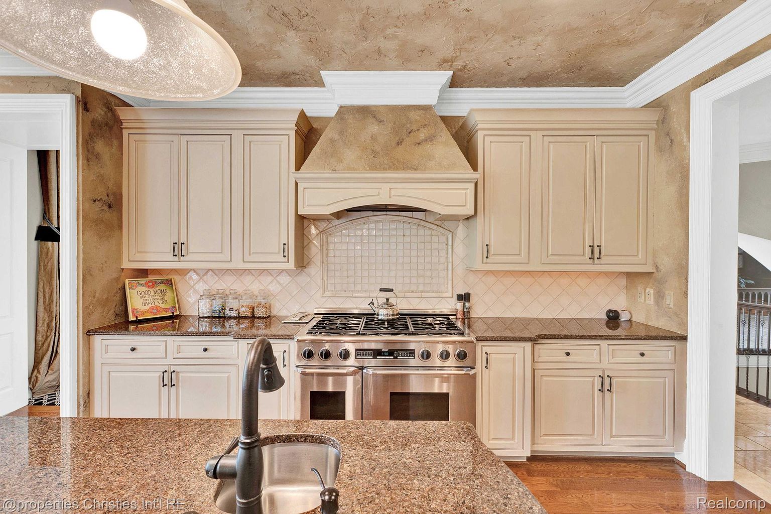 This is a well-appointed kitchen featuring cream-colored cabinetry, dark countertops, and stainless steel appliances. The focal point is a large range with a decorative hood, complemented by a tile backsplash. The kitchen island features a sink and faucet, and the overall design exudes a sense of traditional elegance.